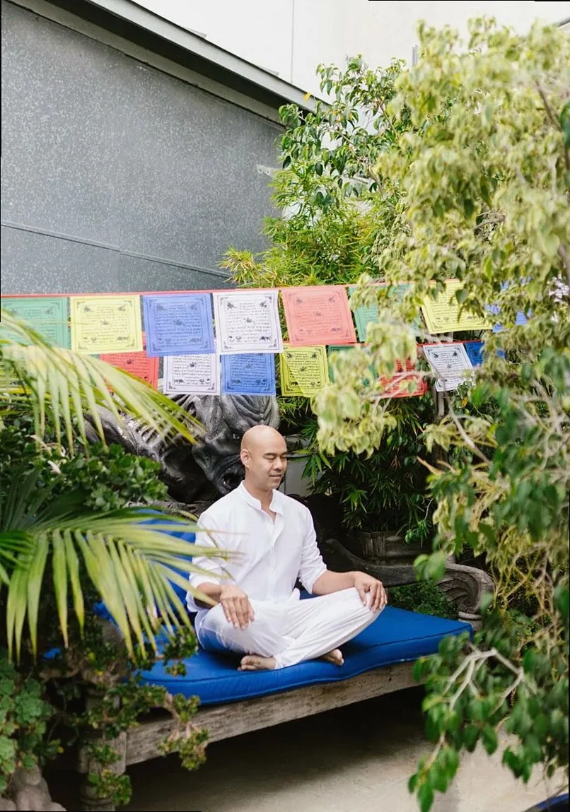 A man in white clothes meditates peacefully outdoors among lush greenery and colorful prayer flags.